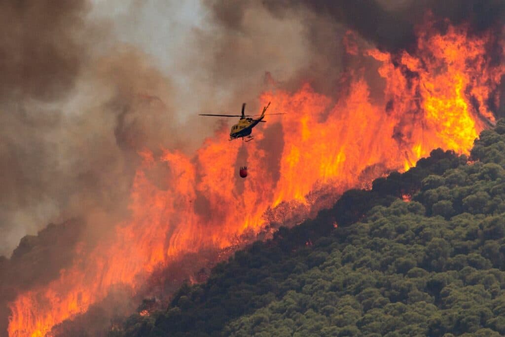 Helicóptero antiincendios sobrevolando un frente de fuego junto a una ladera de monte mediterráneo verde.